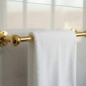 Solid unlacquered brass towel bar with white towel, wall-mounted on marble bathroom wall.
