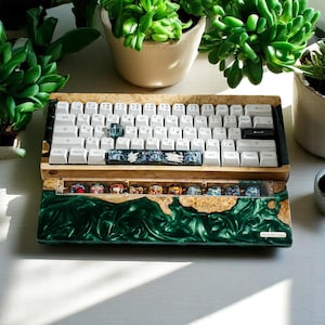 Puede incluir: Un teclado personalizado con teclas blancas y caracteres japoneses. El marco de madera tiene una base de resina verde y una fila superior transparente con teclas coloridas. El teclado está sobre una superficie blanca, rodeado de plantas en macetas.