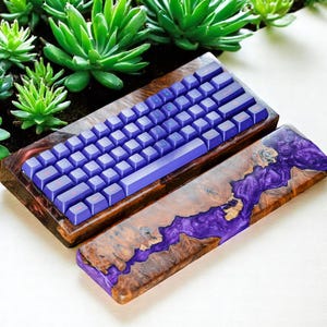 Pode incluir: Um teclado mecânico personalizado com um conjunto de teclas roxas e um apoio para os pulsos a condizer. A base de madeira tem um padrão de burl único com detalhes em resina roxa. O teclado é de tamanho compacto.