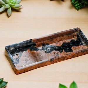 以下が含まれることがあります： 黒、茶色、白の色合いの大理石模様の長方形樹脂トレイ。 トレイには隆起したエッジと小さな真鍮色のアクセントがあります。 トレイは明るい木製の表面に表示され、背景には緑の植物があります。