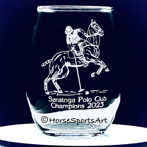 May include: A clear glass with a white etched design of a polo player on a horse. The text "Saratoga Polo Club Champions 2023" and the copyright symbol "©" followed by "HorseSportsArt" are also etched on the glass.