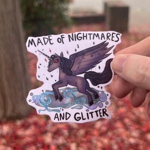 Könnte beinhalten: Aufkleber mit einem dunkelvioletten Einhorn mit schwarzen Flügeln, einem Horn und roten Augen, das auf einer Wolke steht. Der Text "MADE OF NIGHTMARES AND GLITTER" befindet sich über und unter dem Einhorn.