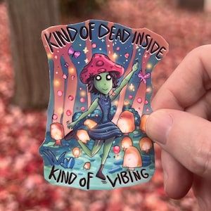 Puede incluir: Una pegatina con una ilustración caprichosa de un personaje con cabeza de hongo con un vestido azul, con el texto "KIND OF DEAD INSIDE" y "KIND OF VIBING". La obra de arte incluye una escena de bosque con luces brillantes y mariposas.