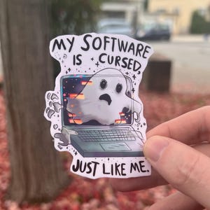 Puede incluir: Una pegatina blanca con el texto "MY SOFTWARE IS CURSED, JUST LIKE ME." La pegatina presenta un fantasma de dibujos animados que emerge de una pantalla de portátil con gráficos con fallos. El fondo está borroso.