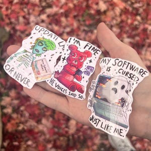 Op de afbeelding: Drie humoristische stickers worden getoond. Eén toont een skelet met de tekst "Update Later or Never". Een andere toont een rode teddybeer met de tekst "I'm Fine". De derde toont een laptop met een spook en de tekst "My Software is Cursed."