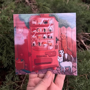 May include: Square print featuring a red sushi vending machine with various sushi types. A tall orange cat is operating the machine, with a panda and white cat nearby. The print has the text "Thank you!" on the vending machine.