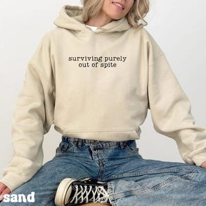 May include: A sand-colored hoodie with the text "surviving purely out of spite" in black. The hoodie has a hood and long sleeves. The person in the image is wearing blue jeans and black and white sneakers.