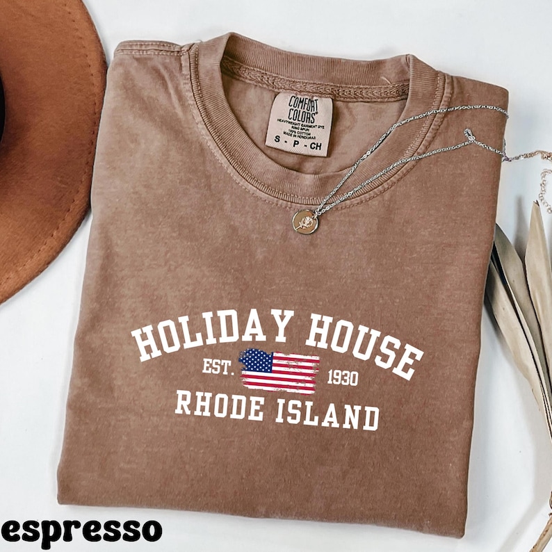 Op de afbeelding: Espresso bruin t-shirt met een witte afbeelding met de tekst "HOLIDAY HOUSE EST. 1930 RHODE ISLAND" en een kleine Amerikaanse vlag.