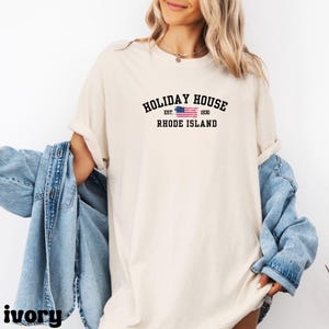 Op de afbeelding: Een wit T-shirt met een zwarte afbeelding waarop staat "HOLIDAY HOUSE EST. 1930 RHODE ISLAND" met een kleine Amerikaanse vlag.