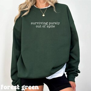 May include: Forest green sweatshirt with the text "surviving purely out of spite" in white. The sweatshirt has long sleeves and a crew neck. The bottom of a white shirt is visible under the sweatshirt.