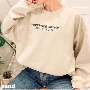 May include: A sand-colored sweatshirt with the phrase "surviving purely out of spite" printed in black. The long-sleeved crewneck sweatshirt is worn with blue jeans. The word "sand" is printed at the bottom of the image.