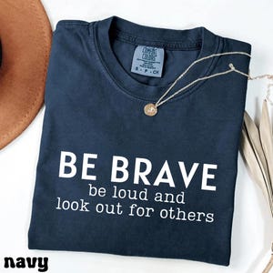 May include: Navy blue t-shirt with the words "BE BRAVE" and the message "be loud and look out for others" printed in white. A gold necklace with a pendant is draped across the shirt.