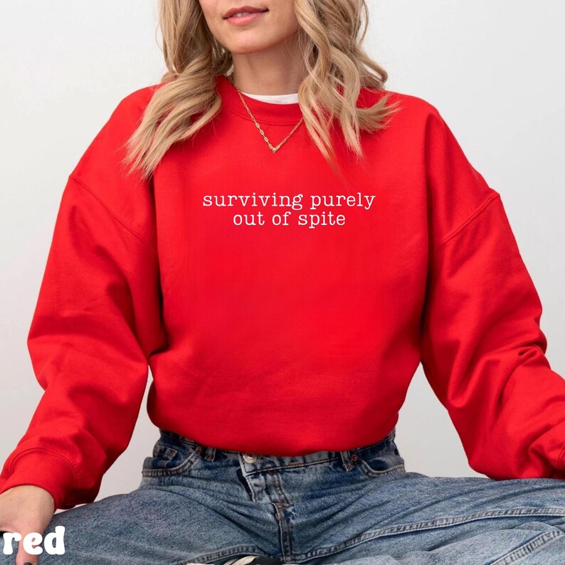 May include: A bright red sweatshirt with the phrase "surviving purely out of spite" printed in white. The sweatshirt is a crew neck style and appears to be made of a soft material. The overall style is casual and comfortable.