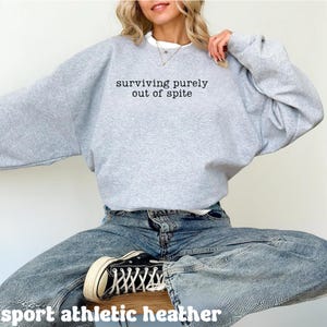 May include: A light gray sweatshirt with the text "surviving purely out of spite" in black. The sweatshirt is paired with blue jeans and black sneakers. The words "sport athletic heather" are at the bottom of the image.