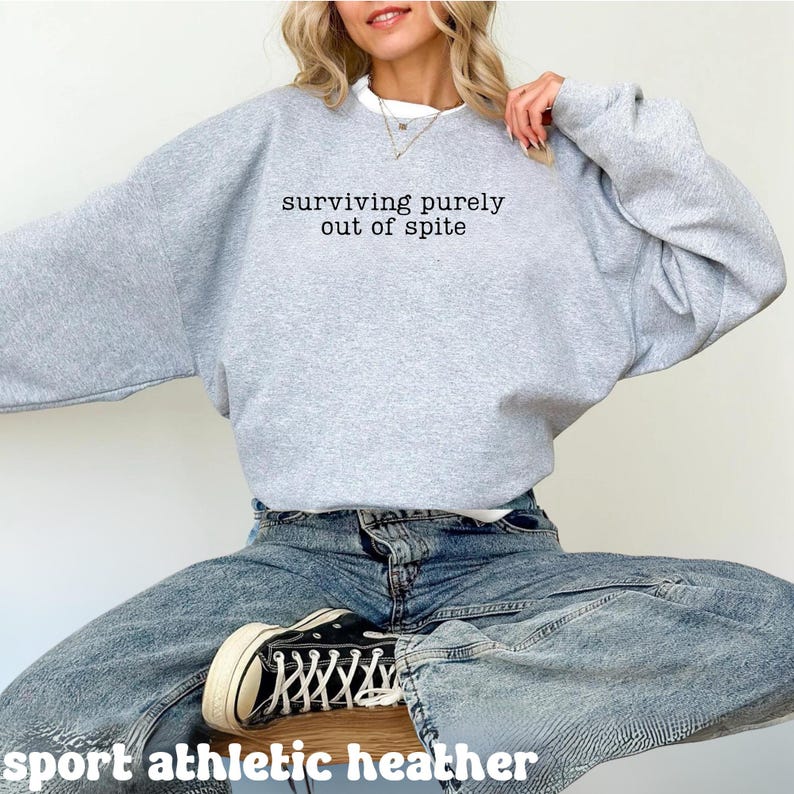 May include: A light gray sweatshirt with the text "surviving purely out of spite" in black. The sweatshirt is paired with blue jeans and black sneakers. The words "sport athletic heather" are at the bottom of the image.