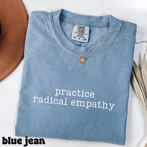May include: A blue jean colored t-shirt with the words "practice radical empathy" in white. A silver necklace with a small pendant rests on the shirt. The shirt is folded and has a tag that says "Comfort Colors".