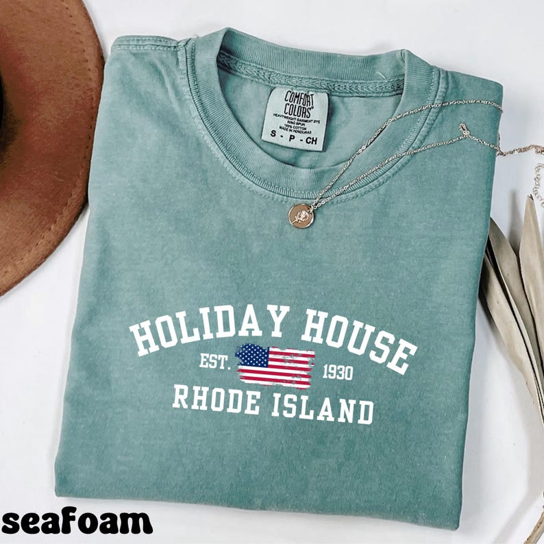 Op de afbeelding: Zeegroen T-shirt met een vintage-stijl grafische afbeelding met de tekst "HOLIDAY HOUSE EST. 1930 RHODE ISLAND" en een kleine Amerikaanse vlag.