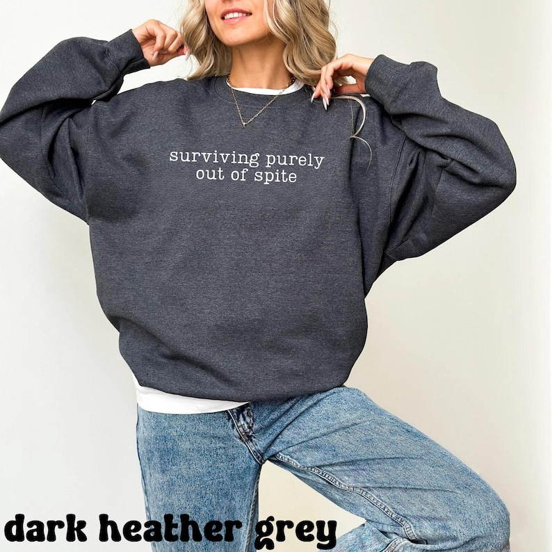 May include: Dark heather grey sweatshirt with the text "surviving purely out of spite" in white. The sweatshirt is a relaxed fit and is paired with blue jeans. The words "dark heather grey" are at the bottom of the image.