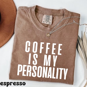 May include: A brown t-shirt with the text "Coffee is my personality" printed in white. The shirt is made of a soft, comfortable material and has a relaxed fit.