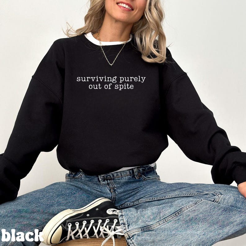 May include: Black sweatshirt with the text "surviving purely out of spite" in white. The sweatshirt has a white collar and is paired with blue jeans and black sneakers.