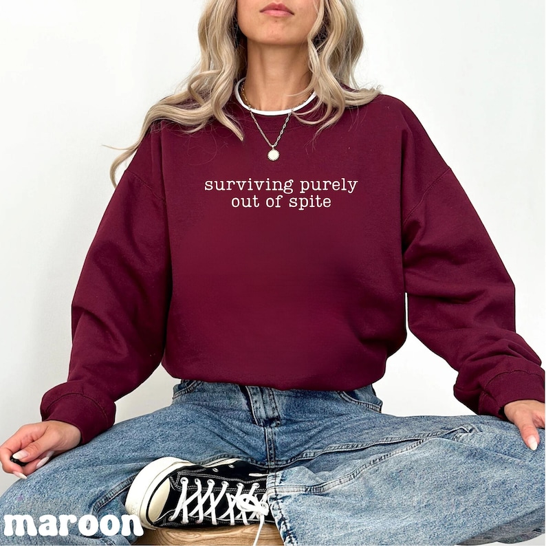 May include: Maroon sweatshirt with the text "surviving purely out of spite" in white. The sweatshirt is a solid color and has long sleeves. The word "maroon" is printed at the bottom left corner of the image.