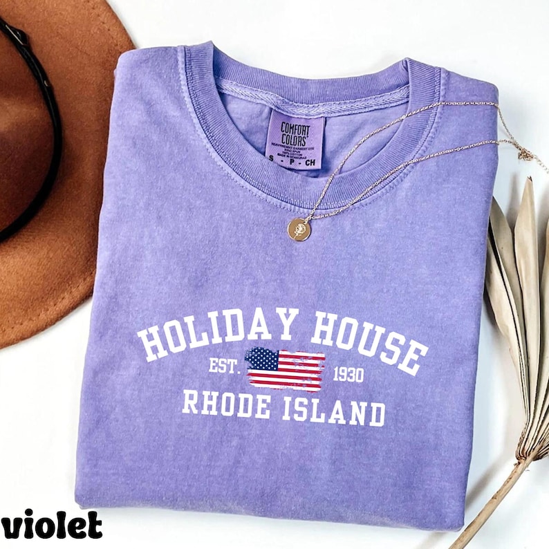 Op de afbeelding: Een paars T-shirt met de tekst "HOLIDAY HOUSE EST. 1930 RHODE ISLAND" in wit. Een kleine Amerikaanse vlag is boven de tekst gedrukt.