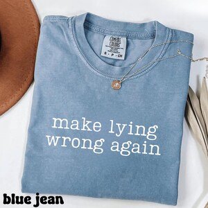 May include: A blue jean colored t-shirt with the phrase "make lying wrong again" printed in white. A silver necklace with a small pendant rests on the shirt. The shirt is folded and placed on a white surface.