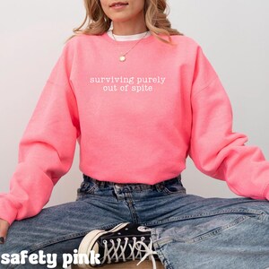 May include: A safety pink sweatshirt with the phrase "surviving purely out of spite" in white lettering. The sweatshirt is a solid color and has long sleeves. The model is wearing blue jeans and black sneakers.