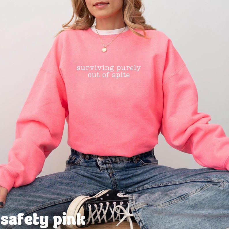 May include: A safety pink sweatshirt with the phrase "surviving purely out of spite" in white lettering. The sweatshirt is a solid color and has long sleeves. The model is wearing blue jeans and black sneakers.
