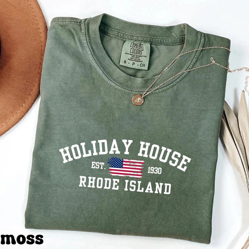 Op de afbeelding: Een groen T-shirt met de tekst "HOLIDAY HOUSE EST. 1930 RHODE ISLAND" in wit. Een kleine Amerikaanse vlag is naast de tekst "EST." gedrukt.