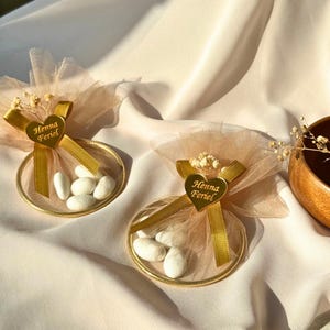 Könnte beinhalten: Zwei goldgerahmte Gastgeschenktüten mit weißen Süßigkeiten, gebunden mit goldenen Bändern und einem herzförmigen Anhänger mit der Aufschrift "Henna Feriel". Die Tüten sind mit beigem Tüll und kleinen Trockenblumen verziert, vor einem weichen, neutralen Hintergrund.