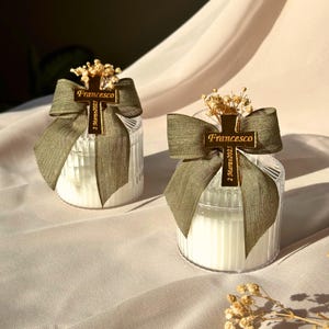 May include: Two clear glass candle holders with white candles inside. Each candle is decorated with a gold cross-shaped charm, a green ribbon, and dried flowers. The charm has the name "Francesco" and the date "2 Marzo 2025" engraved on it.