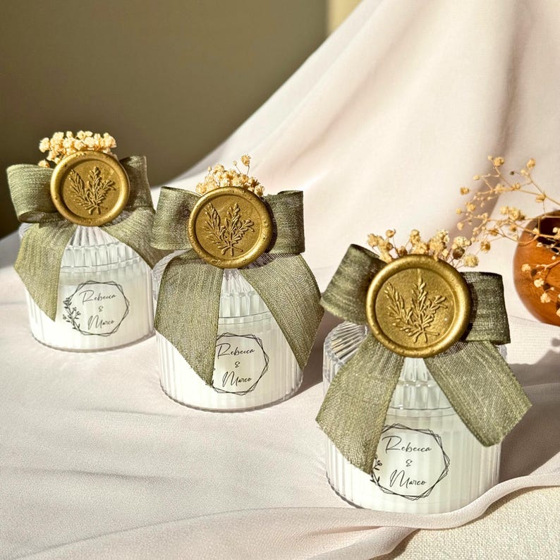Puede incluir: Tres velas blancas de vidrio acanalado con cinta verde oliva y sellos de cera dorados con relieve de hojas. Cada vela tiene la inscripci&oacute;n "Rebecca & Marco". Las velas est&aacute;n decoradas con flores secas.