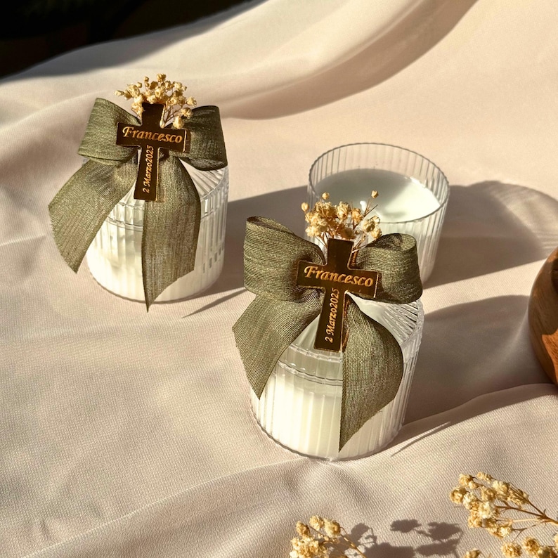May include: Two clear glass candle holders with white candles, adorned with olive green bows and gold crosses. The crosses are inscribed with the name "Francesco" and the date "2 Marzo2025". Dried flowers are placed on top of the candles.