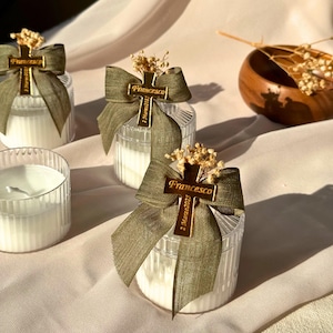 May include: Clear glass candle holders with white candles, adorned with olive green ribbons and gold cross charms. The charms read "Francesco" and "2 Marzo 2023". Dried flowers top each candle, creating an elegant, personalized gift.