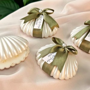 May include: White, pearlised seashell-shaped favour boxes, some with olive green ribbon bows and a pearl accent. The boxes are on a pink fabric surface. Some boxes have tags with handwritten text.