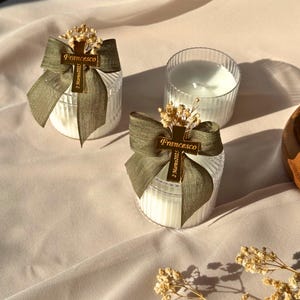 May include: Two white candles in clear, ribbed glass containers, adorned with olive green bows and gold cross charms that read "Francesco" and "2 Marzo 2023." The candles are decorated with small dried flowers, set on a light pink fabric.
