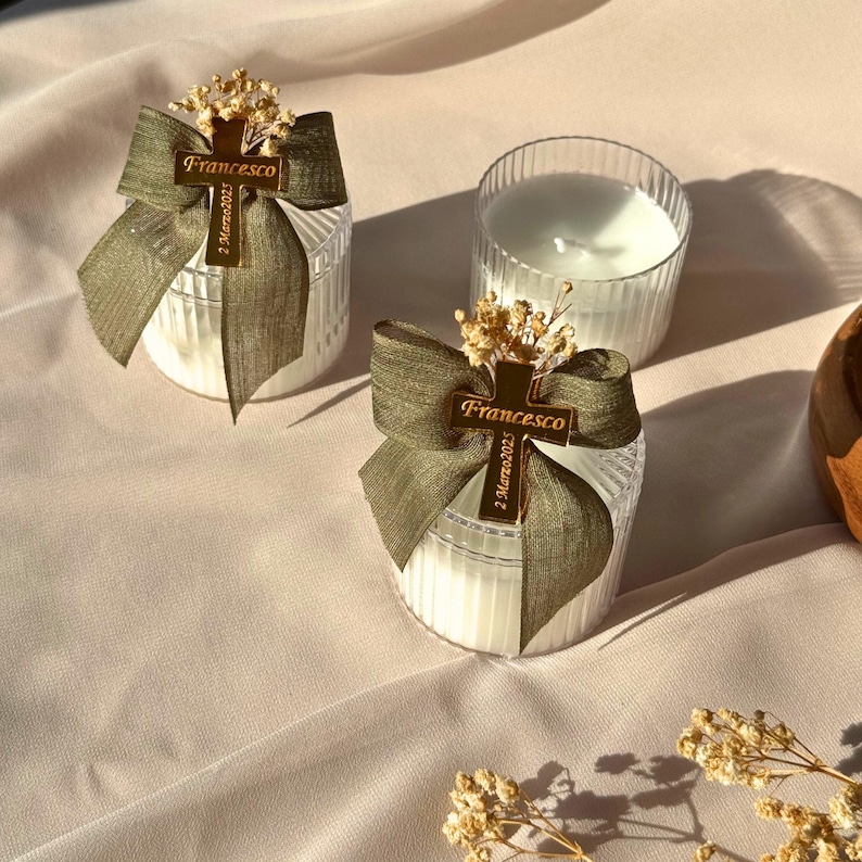 May include: Two white candles in clear, ribbed glass containers, adorned with olive green bows and gold cross charms that read "Francesco" and "2 Marzo 2023." The candles are decorated with small dried flowers, set on a light pink fabric.
