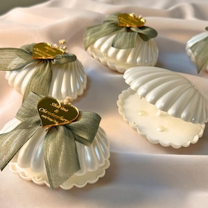 Puede incluir: Contenedores blancos en forma de concha con acabado nacarado, adornados con lazos verde oliva y etiquetas doradas en forma de corazón. Una concha está abierta, revelando pequeñas perlas en su interior. Las etiquetas tienen nombres y una fecha.