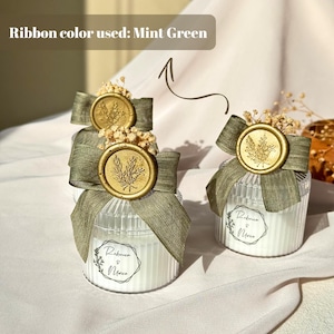 Puede incluir: Frascos de velas de vidrio transparente con cinta verde menta, sellos de cera dorados y acentos florales secos. Cada frasco tiene una etiqueta blanca con los nombres "Rebecca & Marco". El texto "Ribbon color used: Mint Green" es visible.