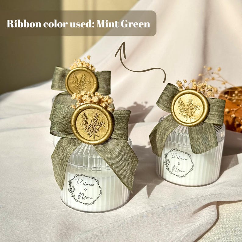 Puede incluir: Frascos de velas de vidrio transparente con cinta verde menta, sellos de cera dorados y acentos florales secos. Cada frasco tiene una etiqueta blanca con los nombres "Rebecca & Marco". El texto "Ribbon color used: Mint Green" es visible.