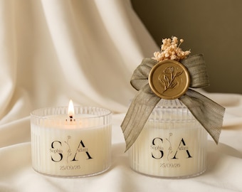 Puede incluir: Dos velas de cristal estriado con cera blanca. Cada vela lleva el texto negro Sophia & Adam y la fecha 25/09/26. Una vela está encendida, mientras que la otra está adornada con una cinta verde, un sello de cera dorado y flores secas.
