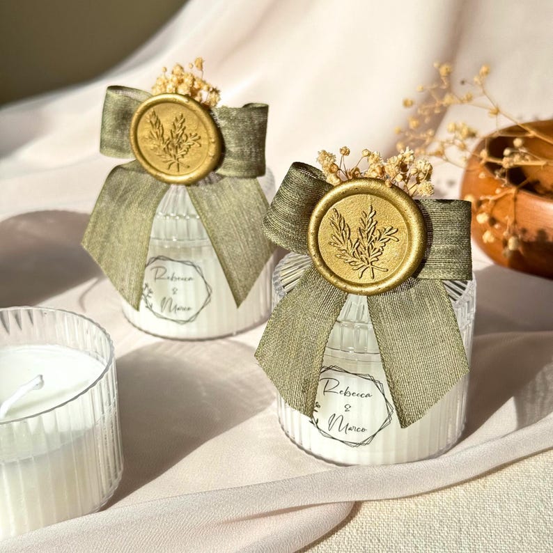 Puede incluir: Dos velas en tarro de cristal con cera blanca, adornadas con cinta verde oliva, sellos de cera dorados y acentos florales secos. Las etiquetas dicen "Rebecca & Marco". Una vela adicional es visible.