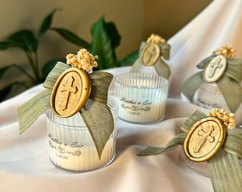 Puede incluir: Candelabros de vidrio transparente con velas blancas, adornados con cintas verde oliva y sellos dorados con cruces. Las velas tienen los nombres "Heather & Eric" y una fecha. Pequeños acentos de flores secas completan el diseño.