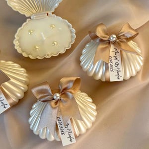 Può includere: Portacandele a forma di conchiglia bianco sporco con dettagli di perle. Ogni portacandele è adornato con un nastro dorato e una perla. Alcuni hanno etichette con testo. Uno è aperto, rivelando una candela all'interno. Il tema generale è elegante e decorativo.