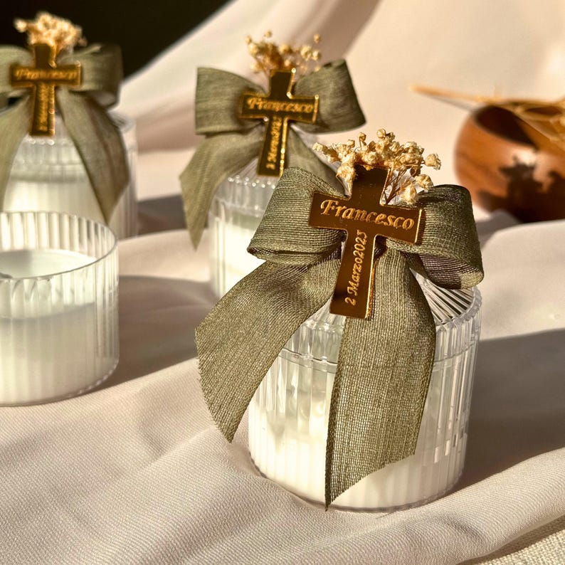 May include: Clear glass candle holders with white candles, adorned with olive green ribbons and gold cross charms. The charms are engraved with the name "Francesco" and the date "2 Marzo 2023". The candles are decorated with dried flowers.
