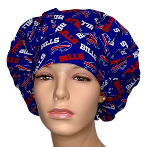 May include: A blue scrub cap featuring a repeating pattern of the Buffalo Bills logo and the word BILLS in red and white text. The cap has a gathered, pleated design with a wide front band and a full, rounded crown that covers the head.