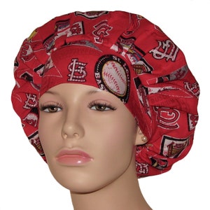 May include: Red and white surgical scrub hat with a St. Louis Cardinals baseball team print. The hat features the team logo, baseballs, and the words "Busch Stadium" and "St. Louis Baseball".