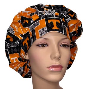 May include: A patterned bouffant scrub cap in orange, black, and gray. The cap features the letter "T" and the words "VOLUNTEERS" and "TENNESSEE" in white. The fabric has a diamond and geometric design.