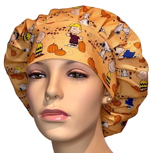 May include: A light orange bouffant surgical scrub cap featuring a repeating pattern of cartoon characters, pumpkins, and autumn leaves. The cap is made of fabric and is designed to cover the hair. The characters include Charlie Brown and Snoopy.